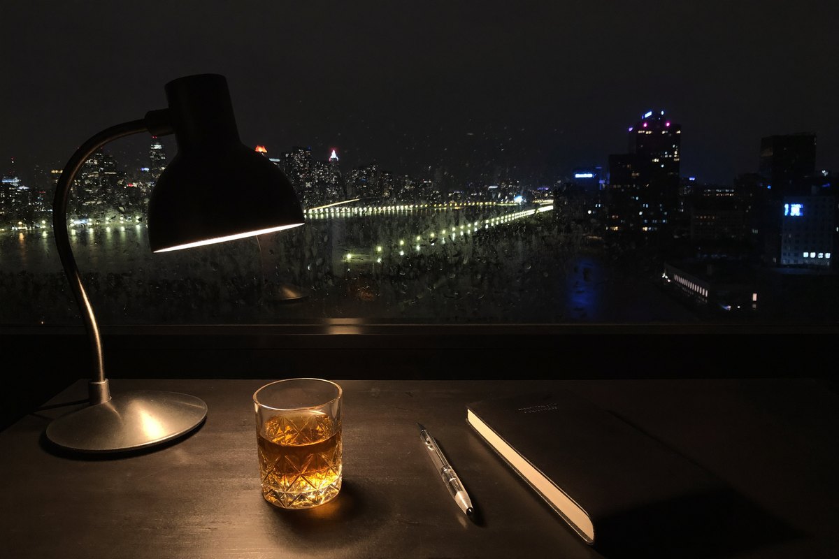 A dark writing desk at night - a single lamp, a tumbler of whiskey, a fountain pen, a cold city skyline through rain-streaked glass.